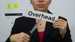 Female office worker or business woman cuts a piece of paper with the word overhead on it as an overhead reduction business concept.