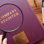 A book with the title Company Register seen through a magnifying glass and calculator next to the book.