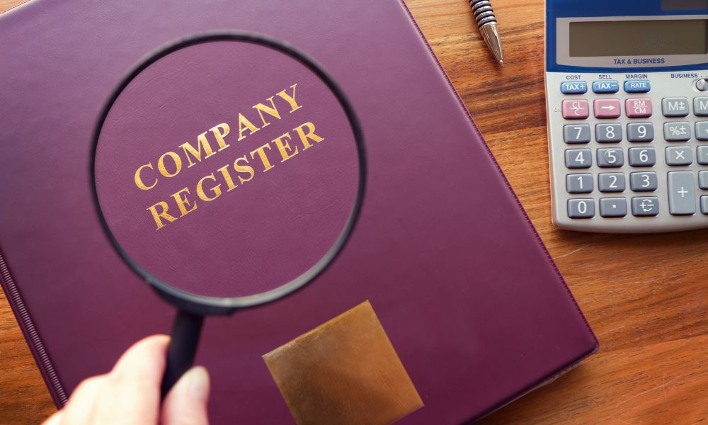 A book with the title Company Register seen through a magnifying glass and calculator next to the book.